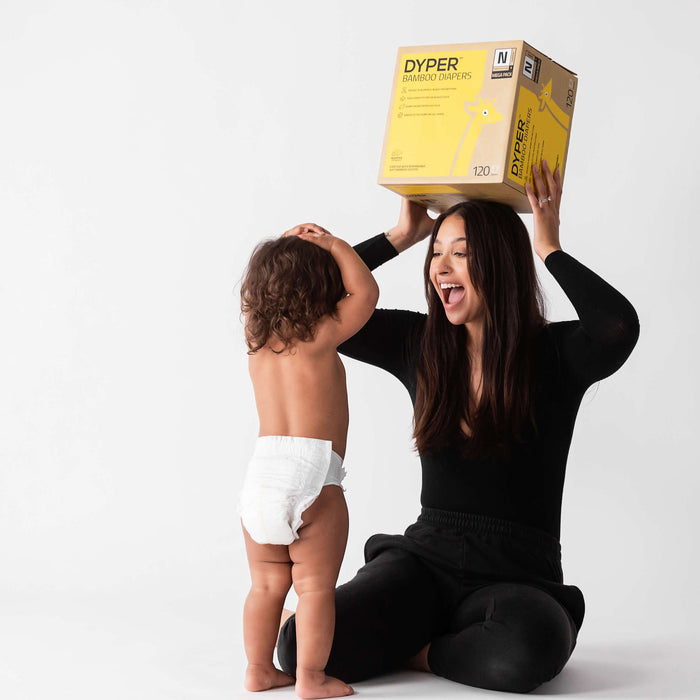 Mom holding a box of disposable diapers over her head next to her baby who is wearing a disposable diaper